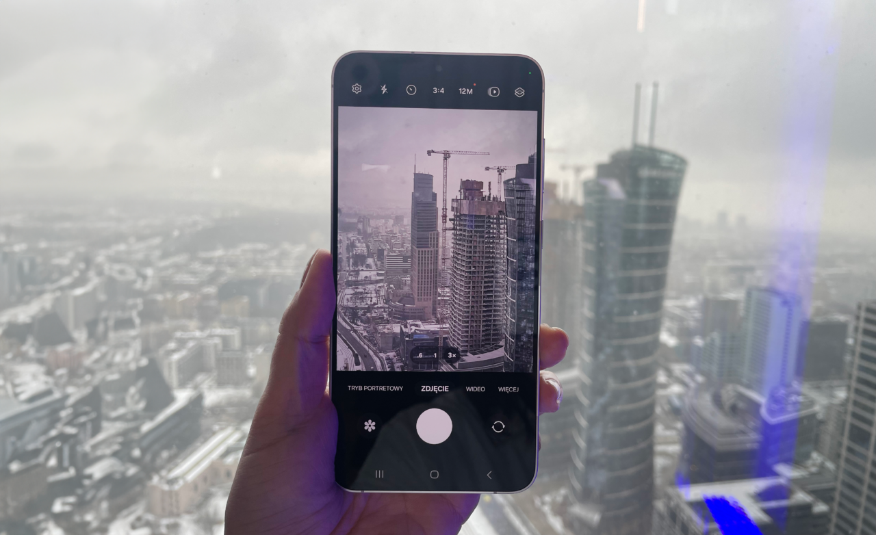Smartfon trzymany w dłoni z wyświetlonym na ekranie widokiem miasta z wysokości, w tle widoczne budynki i rozmyta okolica.