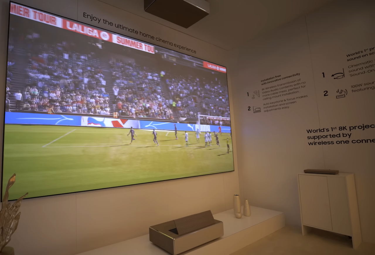 Pokój z dużym ekranem telewizyjnym ,wyświetlającym mecz piłki nożnej, przed którym stoi niska szafka z dekoracyjnymi przedmiotami; na ścianie informacje o projektorze Samsung Premiere 8K.