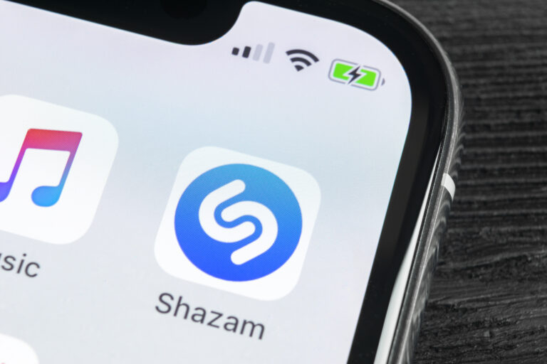 Pierwszy plan ekranu smartfona z widoczną aplikacją Shazam na pulpicie, obok ikon innych aplikacji, na tle czarnej tekstury.