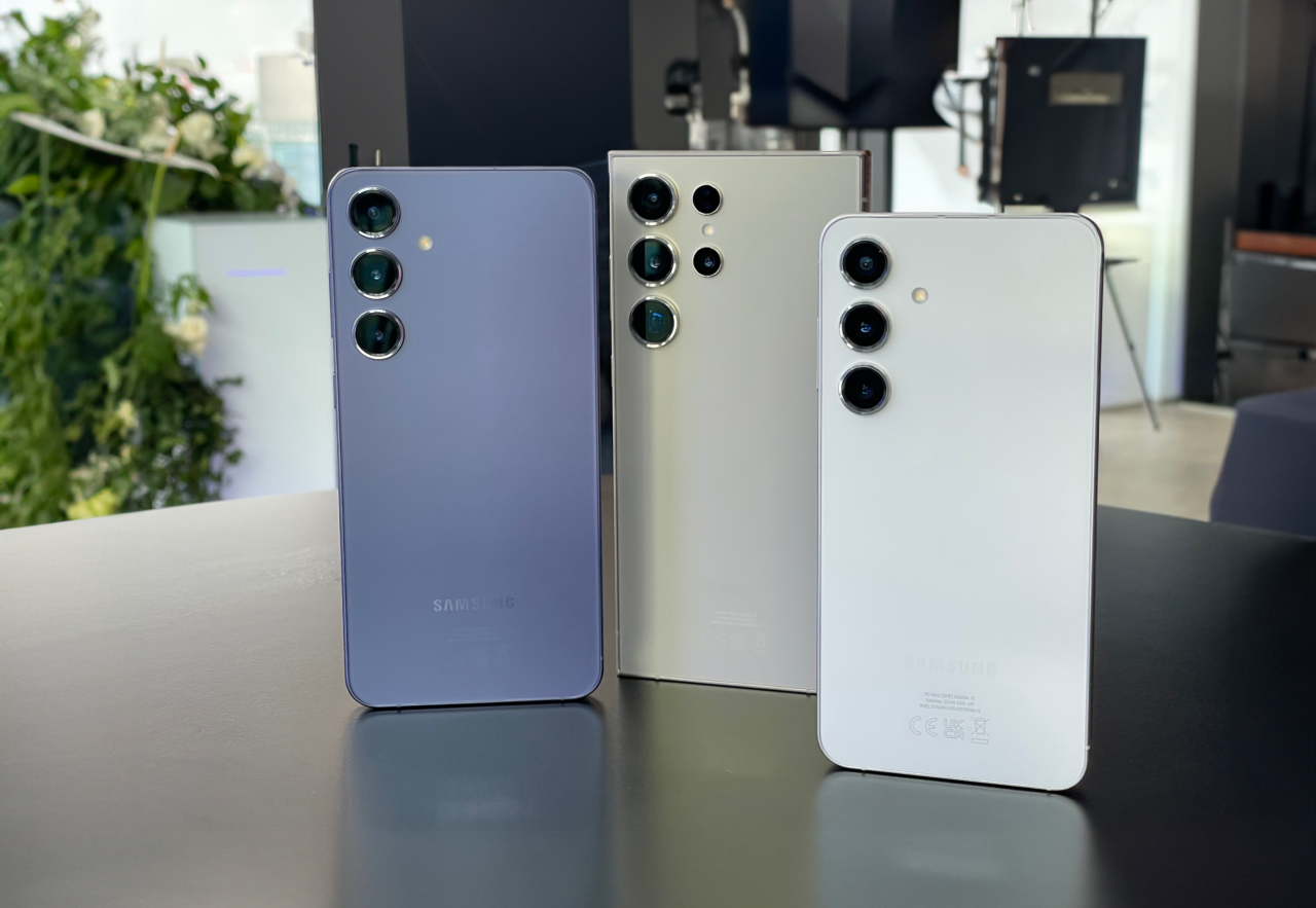Trzy smartfony Samsung galaxy s24 różnych kolorów stojące pionowo na białym blacie z aparatem skierowanym do przodu i widokiem na ich tylne panele z wieloma obiektywami kamery.