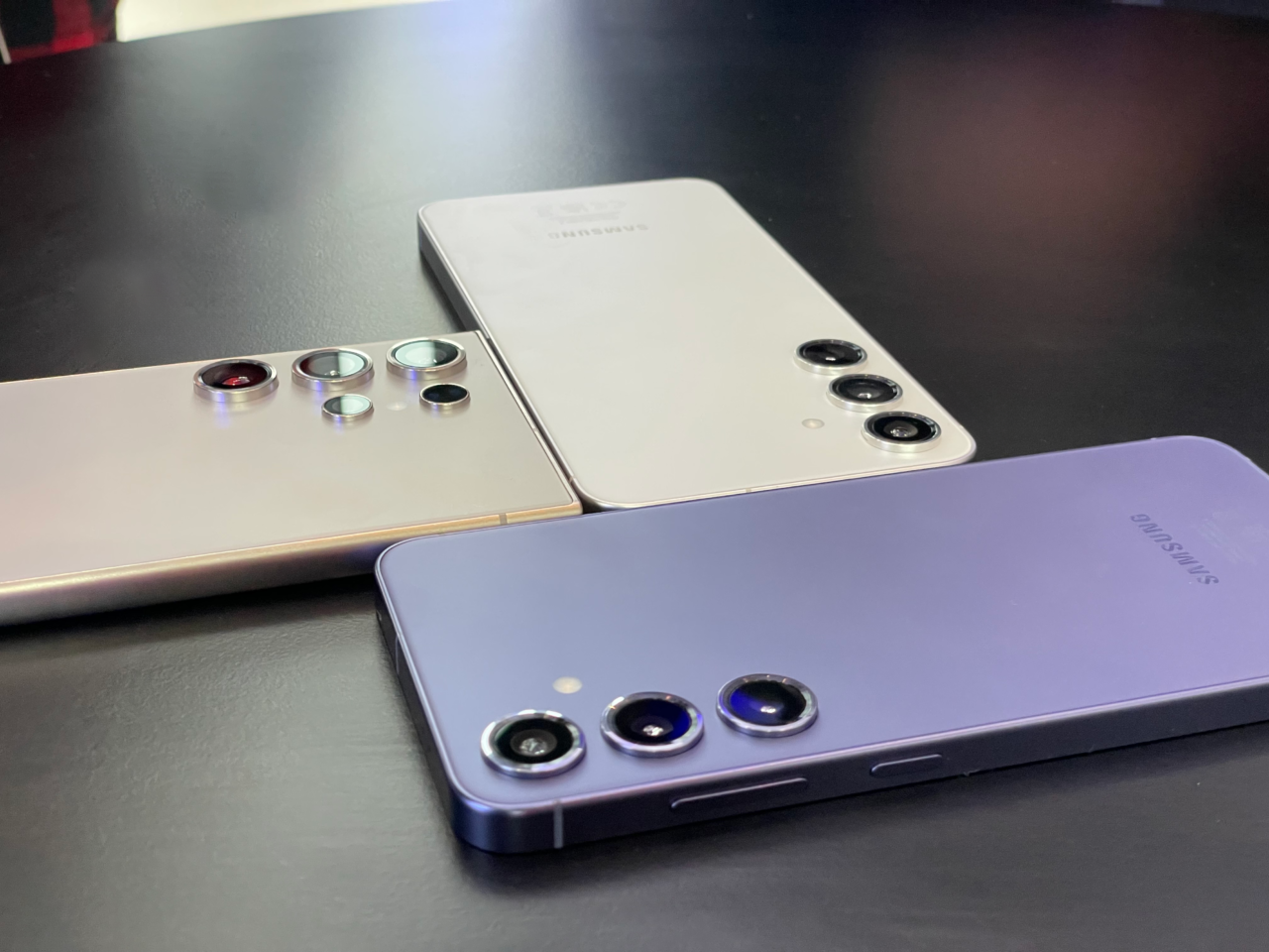 Trzy smartfony Samsung o różnych kolorach i konfiguracjach aparatu umieszczone tyłem na ciemnym biurku.