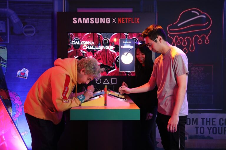 Trzy osoby uczestniczą w wyzwaniu squid game Dalgona Challenge w miejscu promocyjnym Samsung x Netflix.