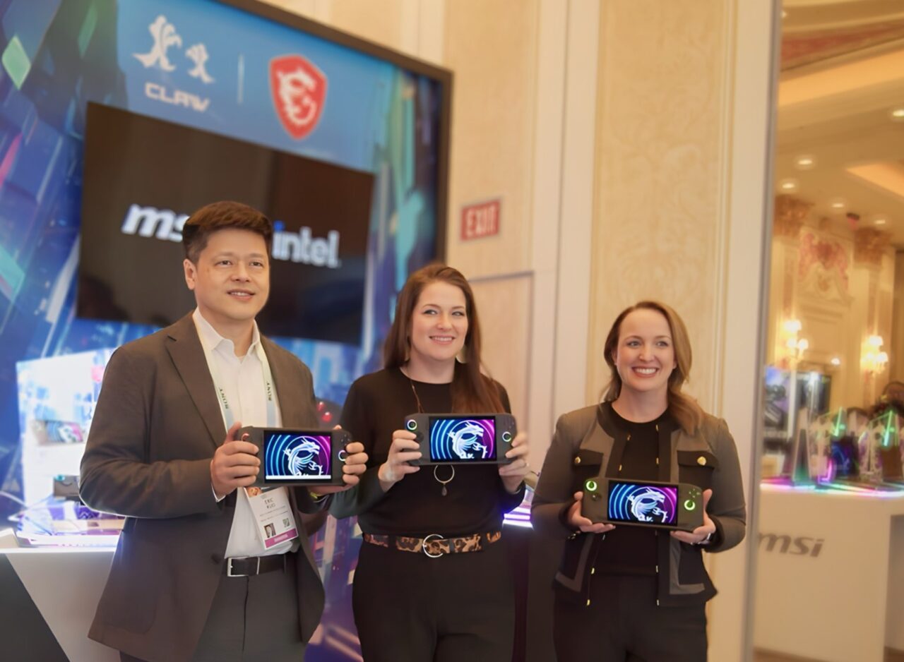 Trzy osoby stojące i prezentujące urządzenia elektroniczne z wizualizacjami na ekranach, na tle wystawienniczego stoiska z logo firm technologicznych.