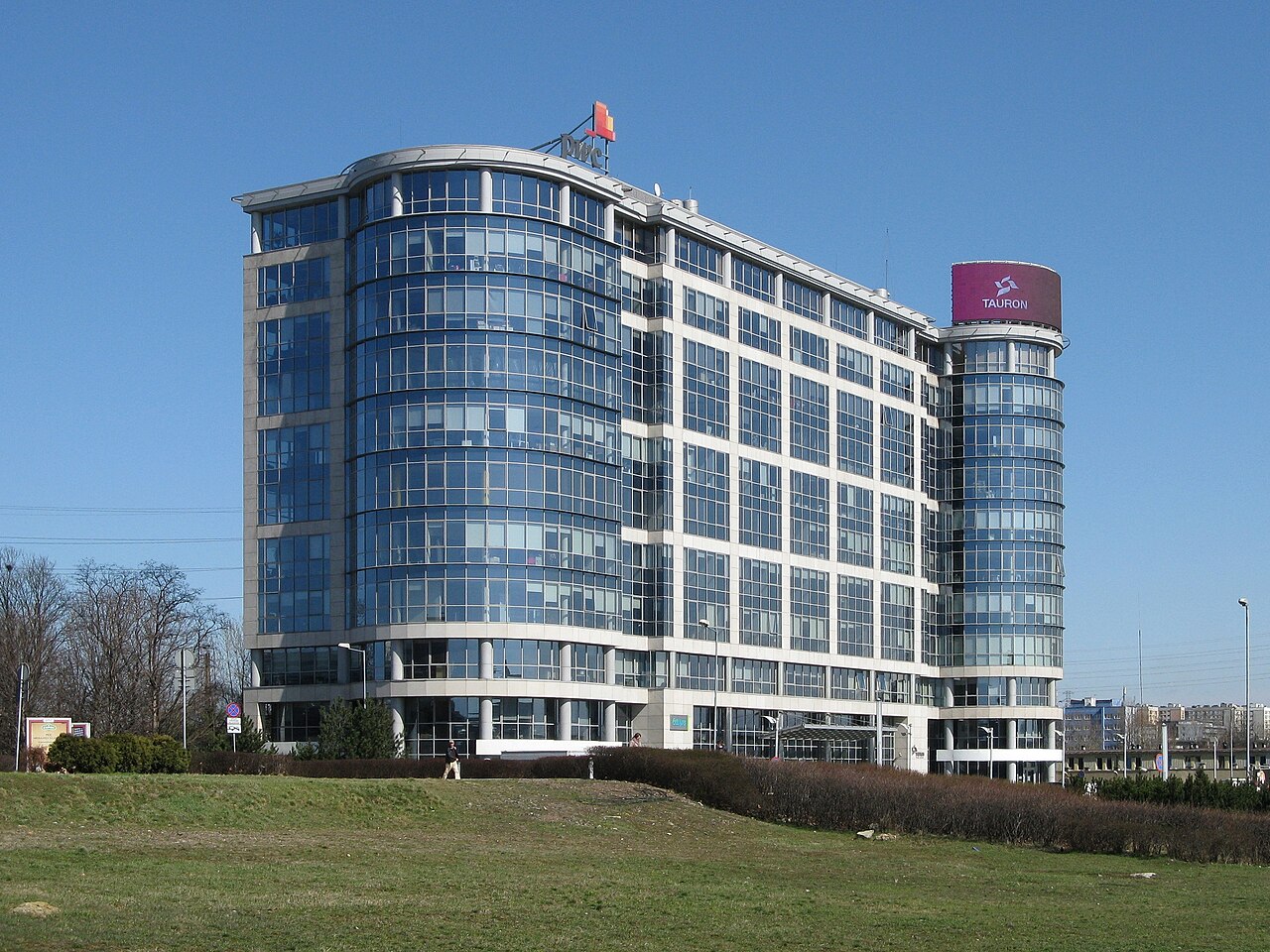 Widok na nowoczesny budynek biurowy z szybami odblaskowymi i zaokrąglonymi narożnikami pod błękitnym niebem. Na dachu budynek ma umieszczony napis "TAURON".