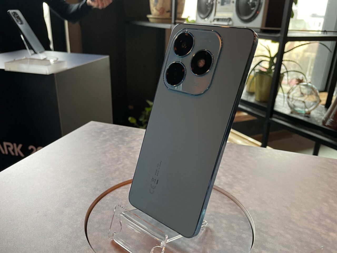 Smartfon tecno spark 20 koloru srebrnego z potrójnym aparatem fotograficznym, wystawiony na stojaku, w jasno oświetlonym wnętrzu.