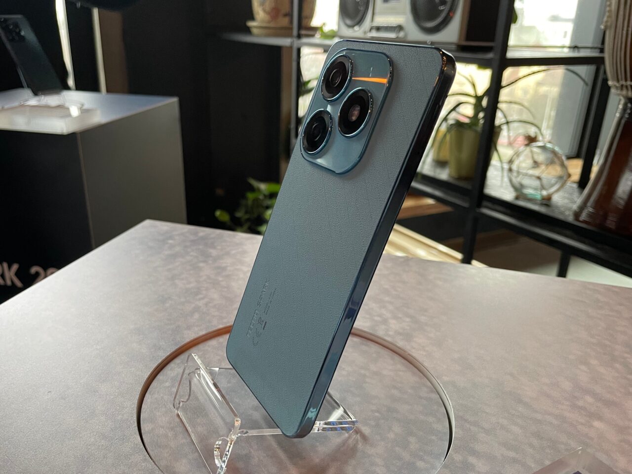 Smartfon tecno spark 20 w niebieskim etui stoi na przezroczystym stojaku, z tyłu widać potrójny aparat fotograficzny.