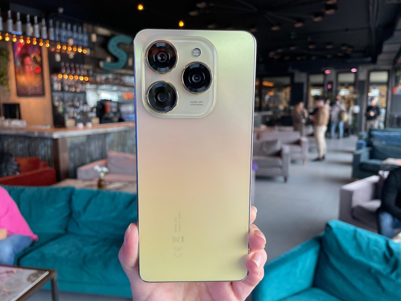 Osoba trzymająca smartfon tecno spark 20 pro z potrójnym aparatem i gradientem kolorów od złotego do białego na tle rozmytego wnętrza kawiarni.
