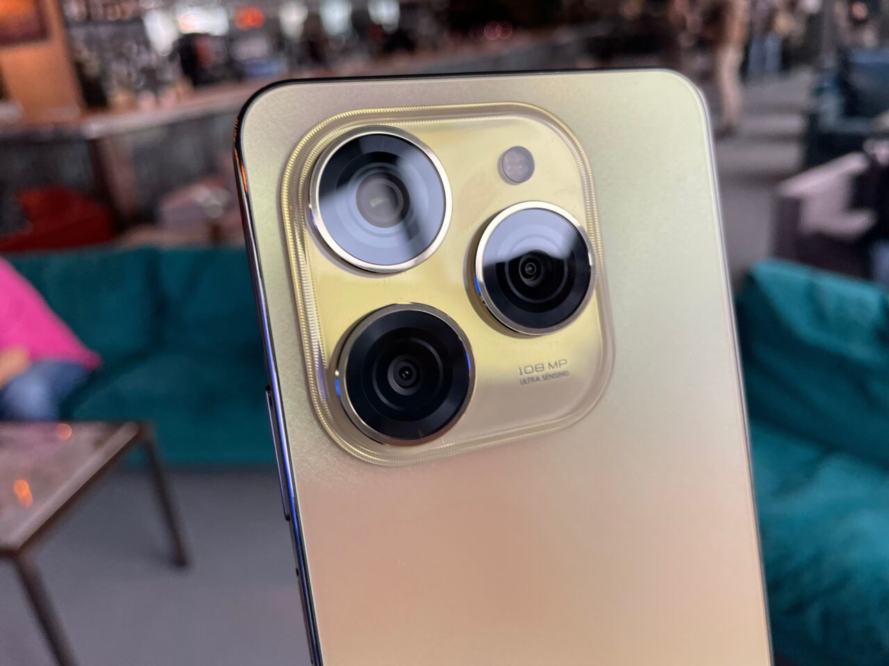 Tylny panel smartfona tecno spark 20 pro w złotym kolorze z trzema obiektywami kamery i etykietą "108 MP ULTRA SENSING" na tle rozmytego wnętrza.