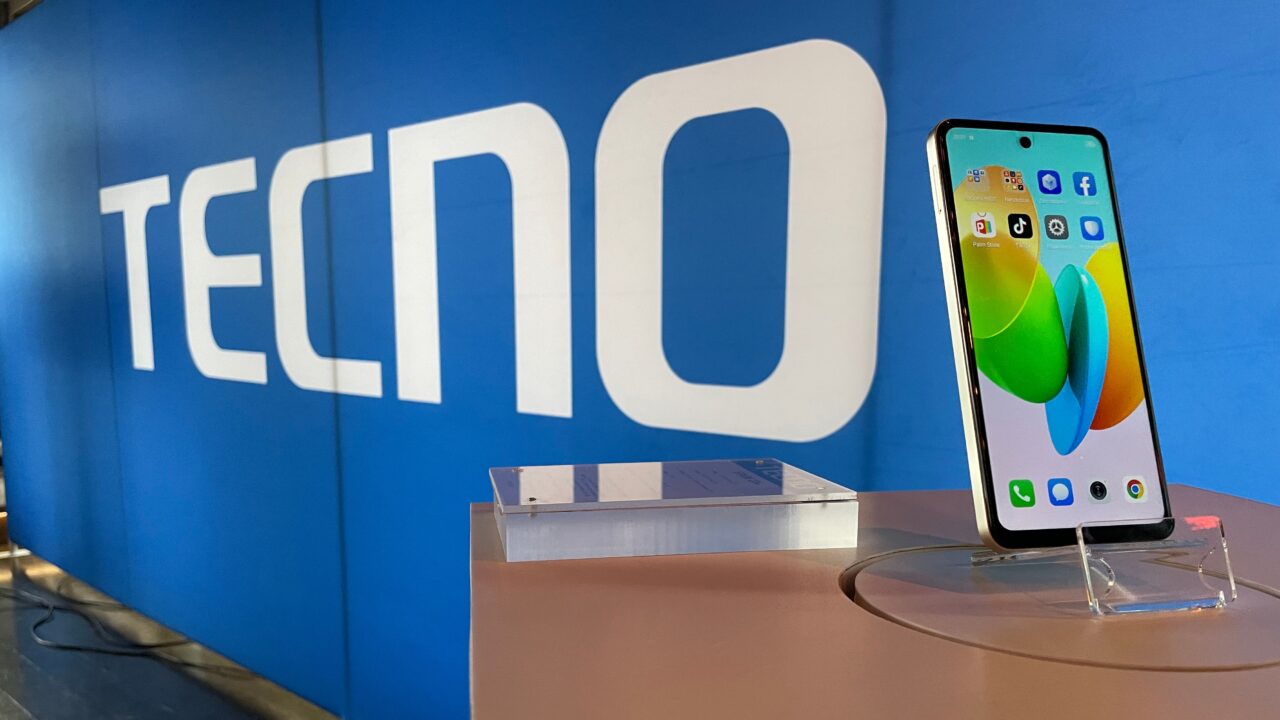 Smartfon TECNO Spark 20 PRO wystawiony na stojaku z logo producenta w tle na ścianie.