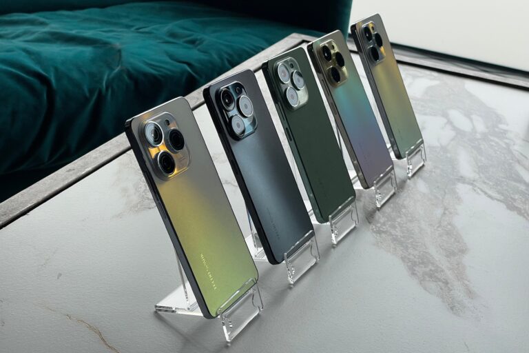 Pięć smartfonów różnych kolorów ustawionych pionowo na przezroczystych stojakach na marmurowym blacie, z zieloną pościelą w tle.