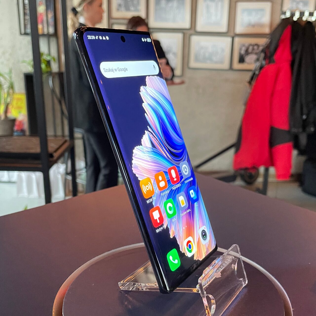 Smartfon tecno spark 20 pro plus umieszczony na przezroczystym stojaku z widocznym wyświetlaczem pokazującym kolorową tapetę i ikony aplikacji, na tle rozmytego wnętrza.