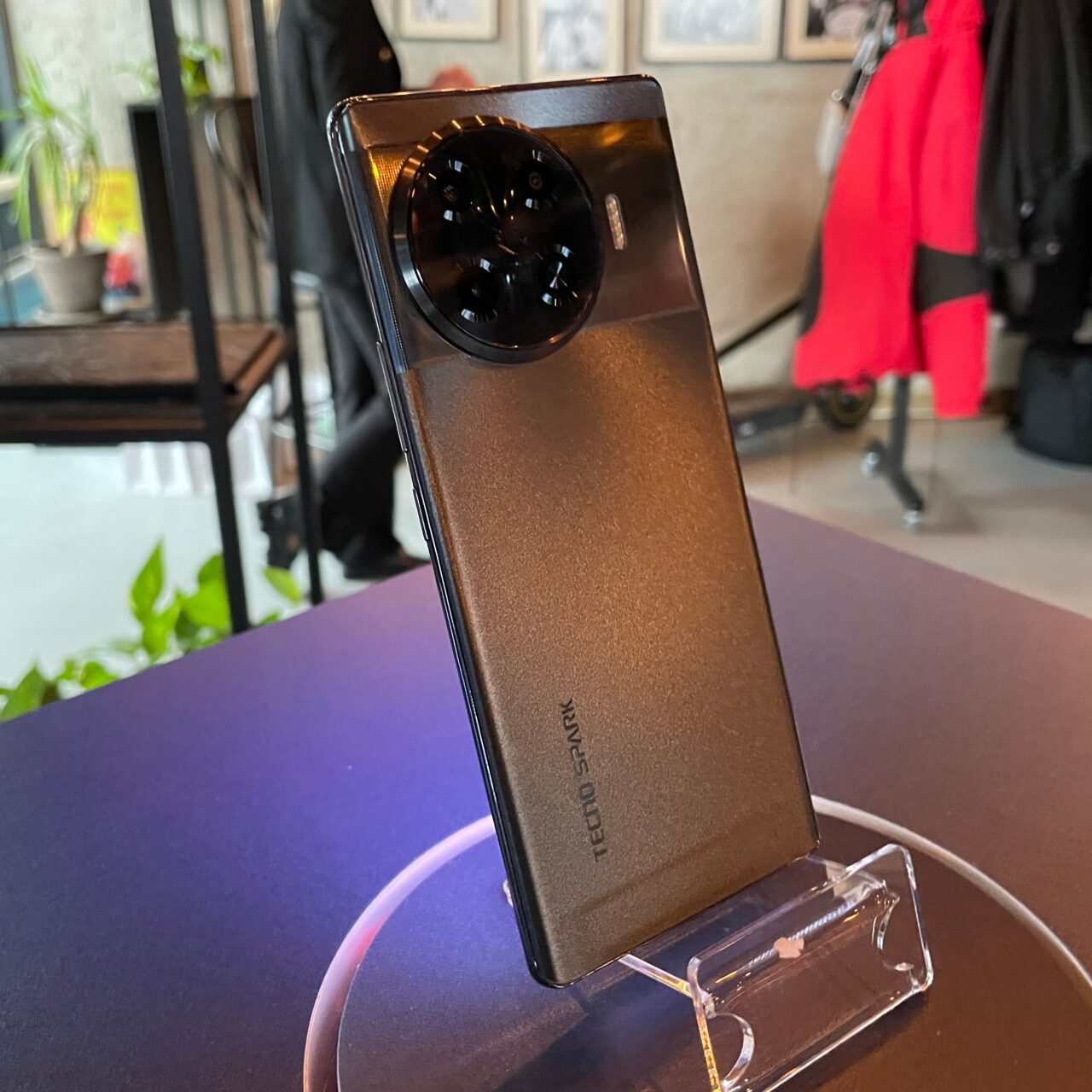 Czarny smartfon tecno spark 20 pro plus z nietypowym okrągłym modułem aparatu umieszczony pionowo na przezroczystym stojaku, w tle wnętrze z roślinami i czerwoną kurtką.