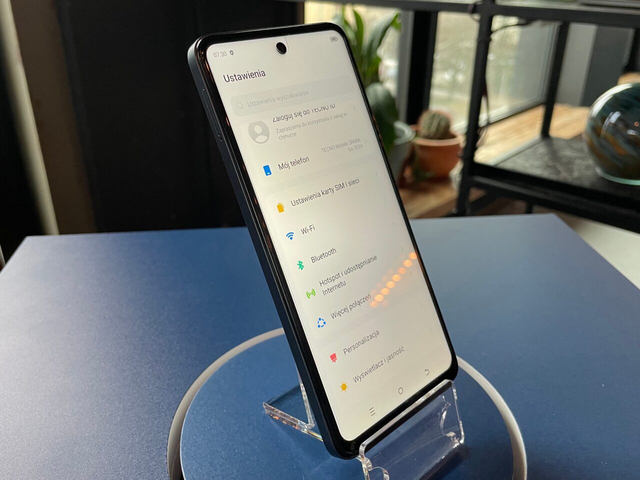 tecno spark go 2024. Smartfon ustawiony pionowo na przezroczystym stojaku, wyświetlający menu ustawień w języku polskim z zaznaczoną opcją baterii i wyświetlania, na tle wnętrza.