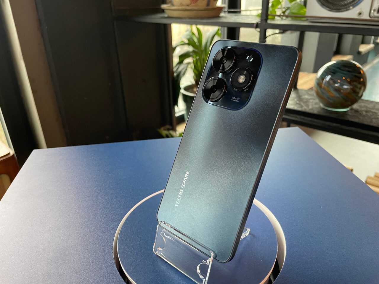 tecno spark go 2024. Smartfon marki Tecno Spark stojący na plastikowym stojaku umieszczonym na niebieskim blacie, z widocznym tylnym aparatem fotograficznym i logo producenta.