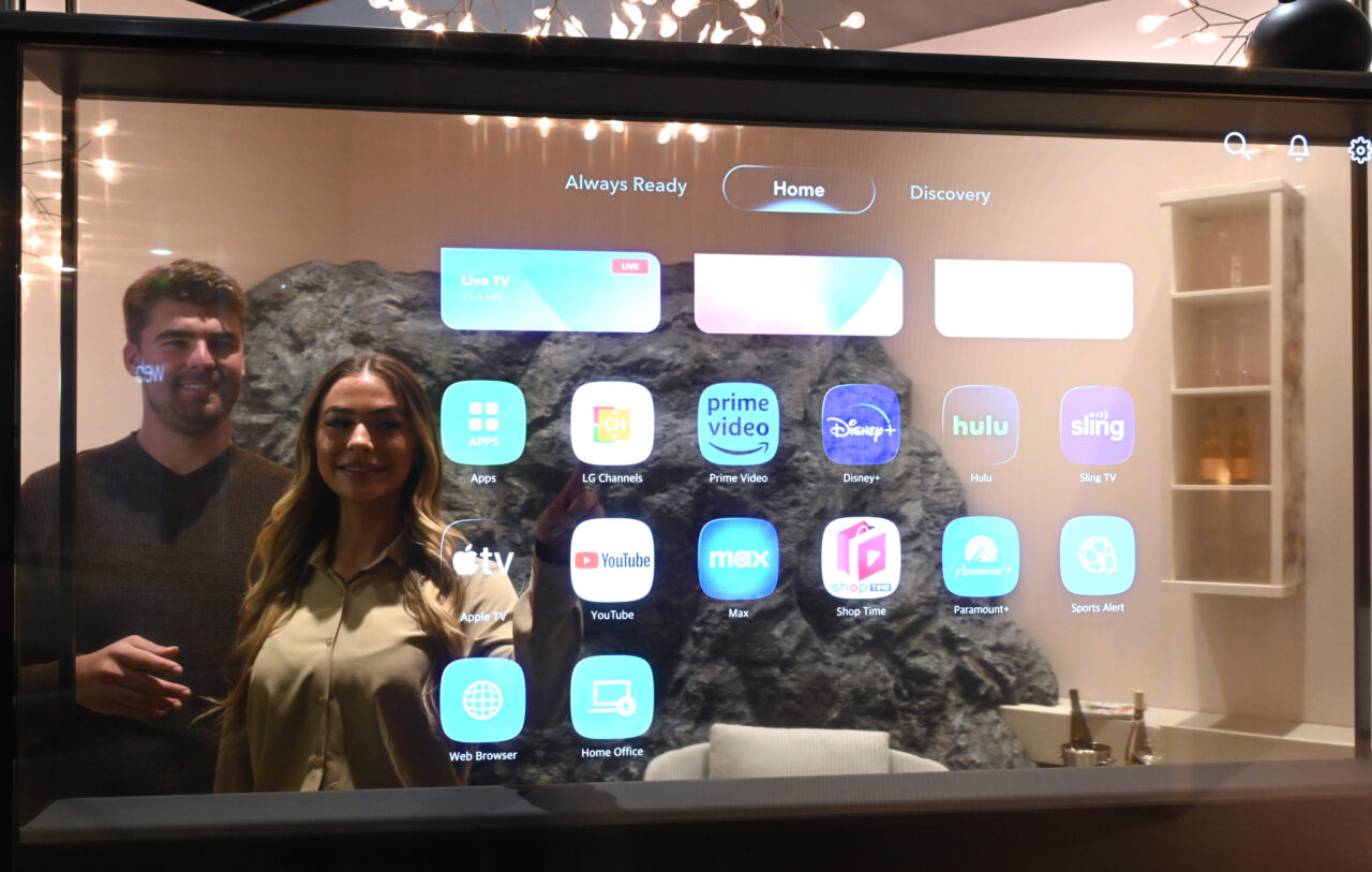 Mężczyzna i kobieta stoją przed telewizor LG Signature OLED T z wyświetlonym interfejsem użytkownika zawierającym aplikacje takie jak Live TV, Prime Video, Disney+, Hulu i inne.