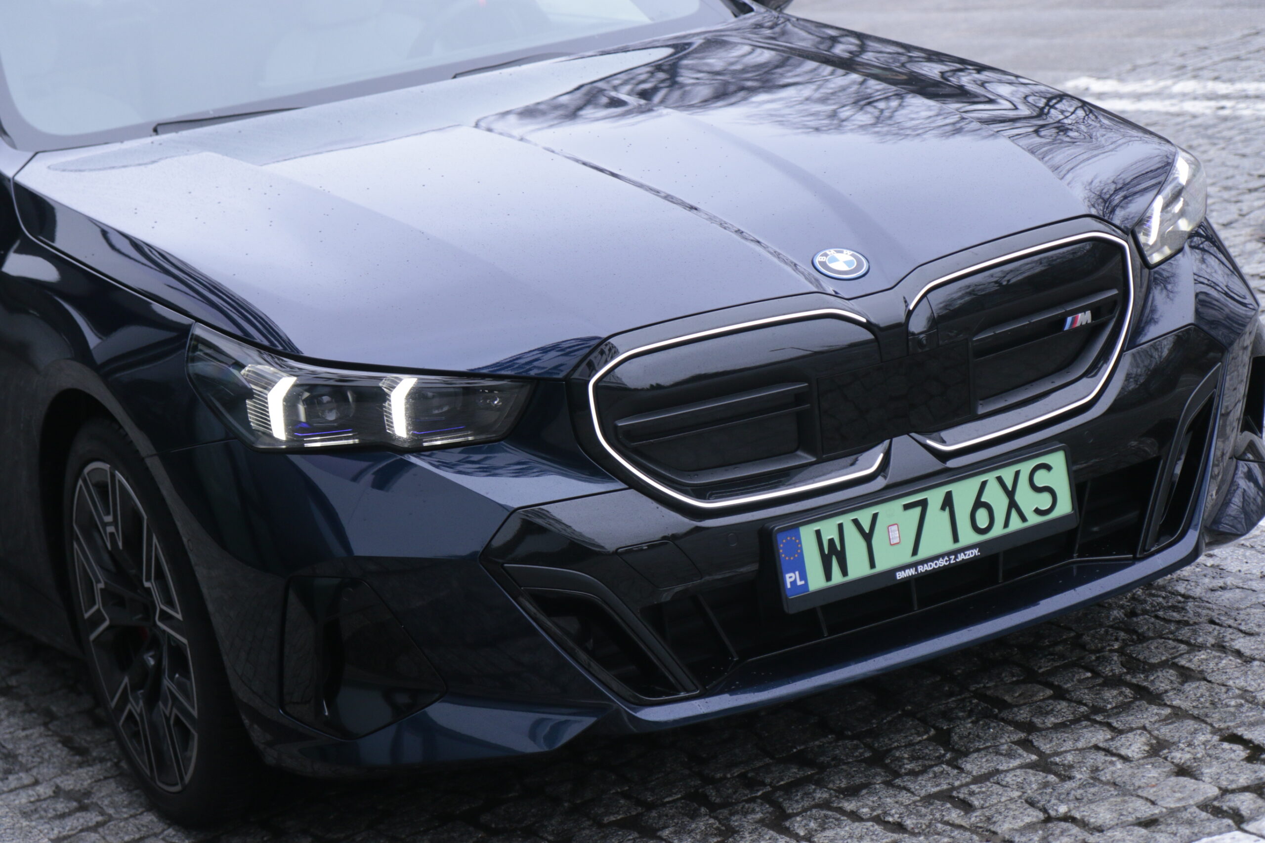 Częściowe zbliżenie na przód testowanego, czarnego samochodu BMW i5 z widocznym logo, oznaczeniem modelu M, reflektorami LED i tablicą rejestracyjną z Polski.