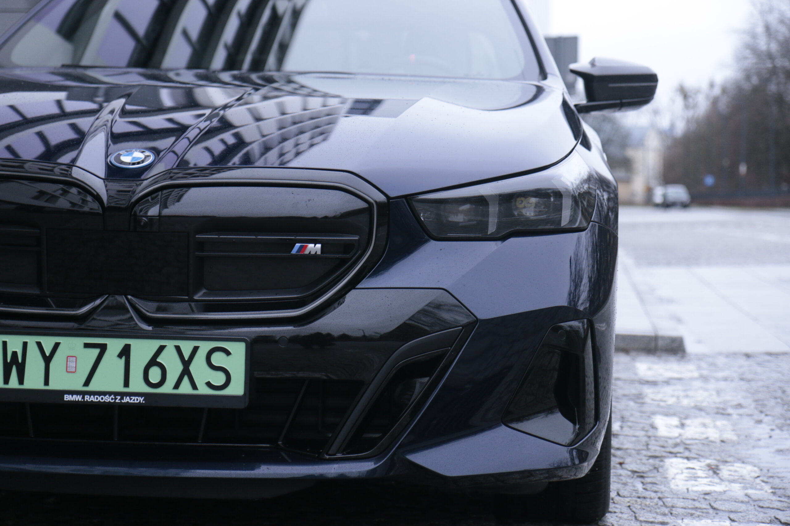 Część przednia czarnego, testowanego samochodu BMW i5 m60 xDrive z emblematem M, zaparkowanego na bruku, z widoczną rejestracją i reflektorami.