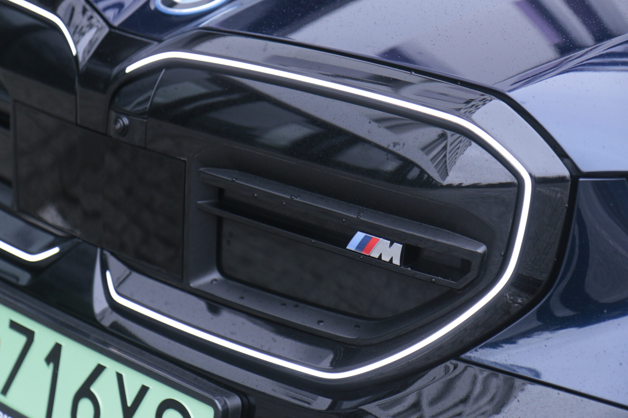 Zdjęcie logo BMW M na przedniej kratce testowanego samochodu i5 M60 xDrive z charakterystycznymi kolorami niebieskim, czerwonym i niebiesko-fioletowym na czarnej tarczy z białą literą „M”.