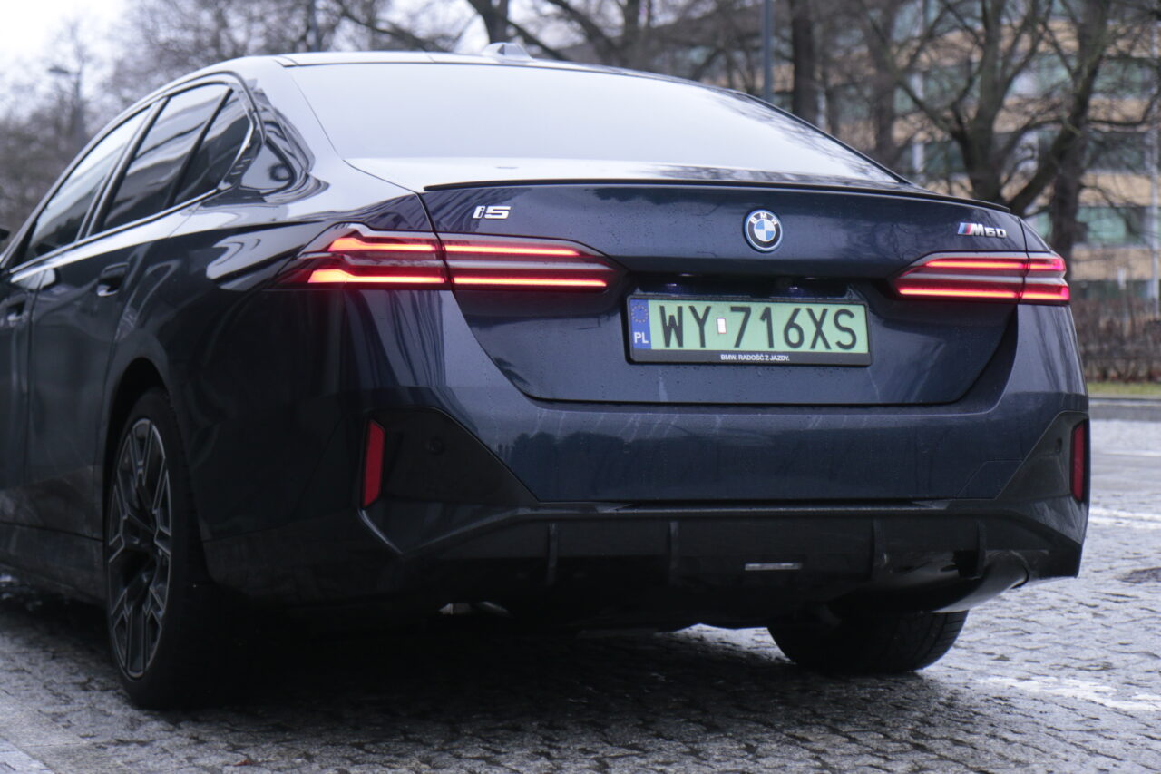 Ciemnogranatowy samochód marki BMW model i5 M50, widziany od tyłu na mokrej nawierzchni, z polskimi tablicami rejestracyjnymi.
