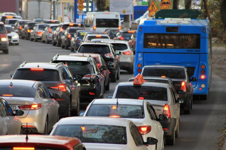 Korek uliczny w dzień, auta ustawione w długim rzędzie, w tle niebieski autobus miejski i znaki drogowe.