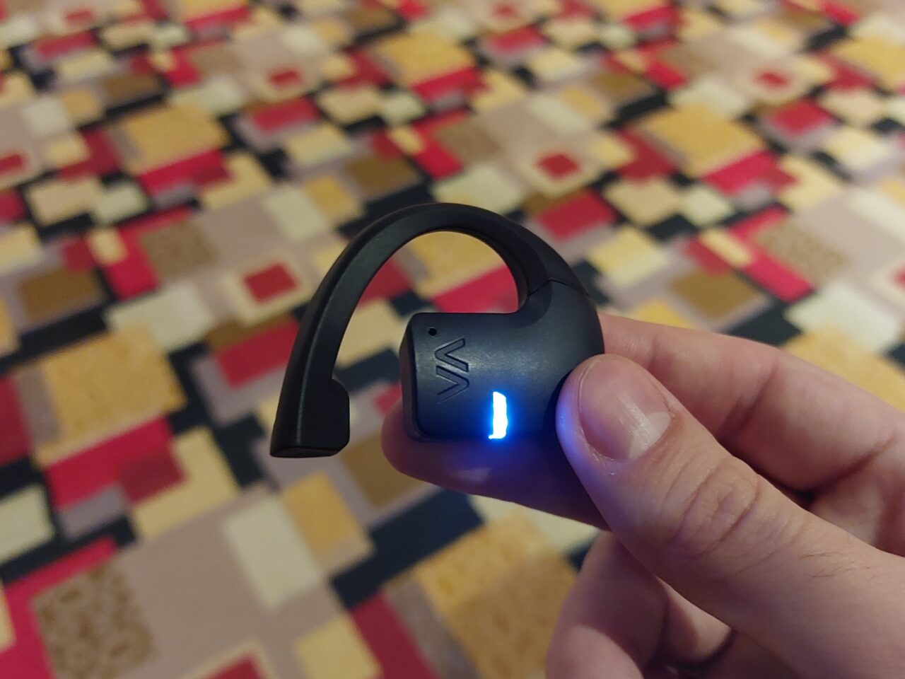 Czarny bezprzewodowy zestaw słuchawkowy Bluetooth trzymany w dłoni na tle wzorzystej powierzchni.