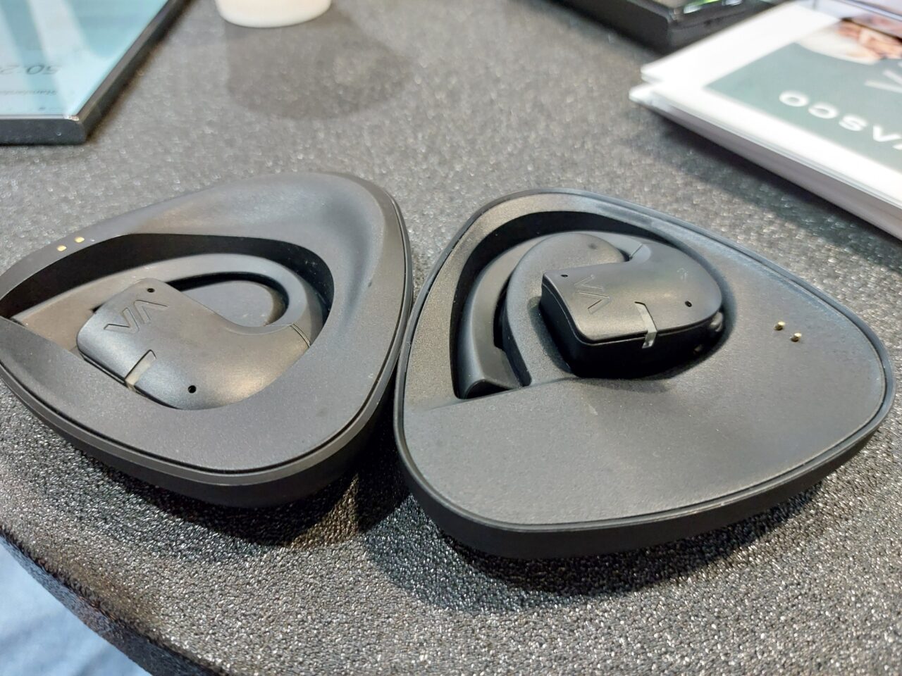 Otwarte czarne etui z bezprzewodowymi słuchawkami dousznymi Vasco Translator E1 leżące na szarym biurku.