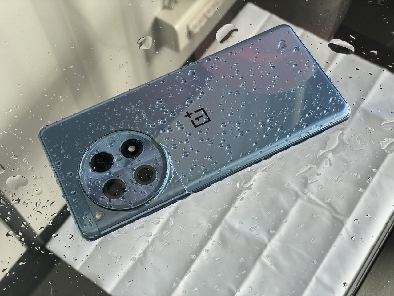 Niebieski smartfon z potrójnym aparatem leży na mokrej, błyszczącej powierzchni z kroplami wody.