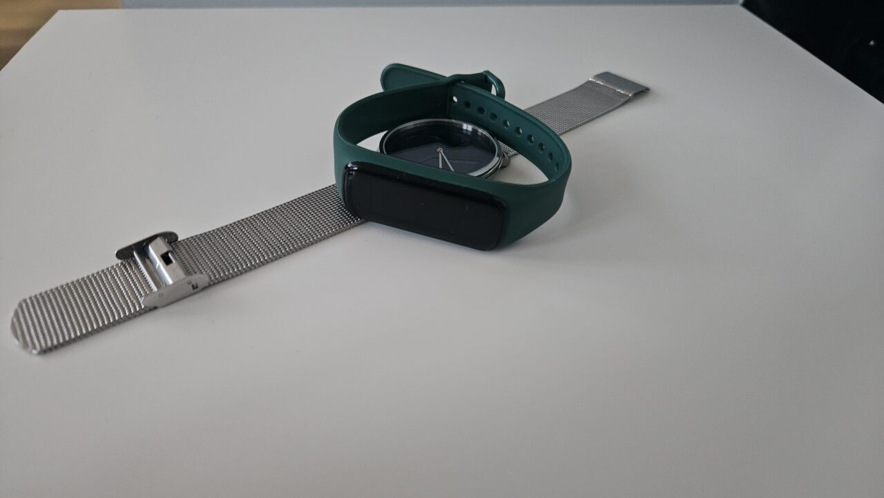 srebrny zegarek z niebieską tarczą i zielona opaska typu smartband z zielonym paskiem leżące na białym stoliku