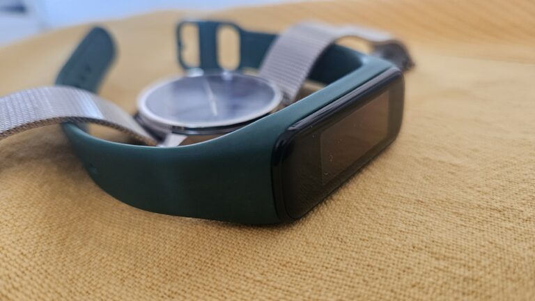 opaska typu smartband Samsung Galaxy Fit 2 z wyłączonym ekranem i srebrny zegarek z niebieską tarczą leżące na żółtym materiale