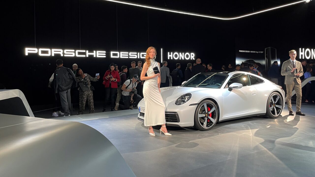 Kobieta w białej sukience stoi obok białego samochodu Porsche na tle ściany z napisem "Porsche Design" w jasno oświetlonym pomieszczeniu z innymi ludźmi w tle.