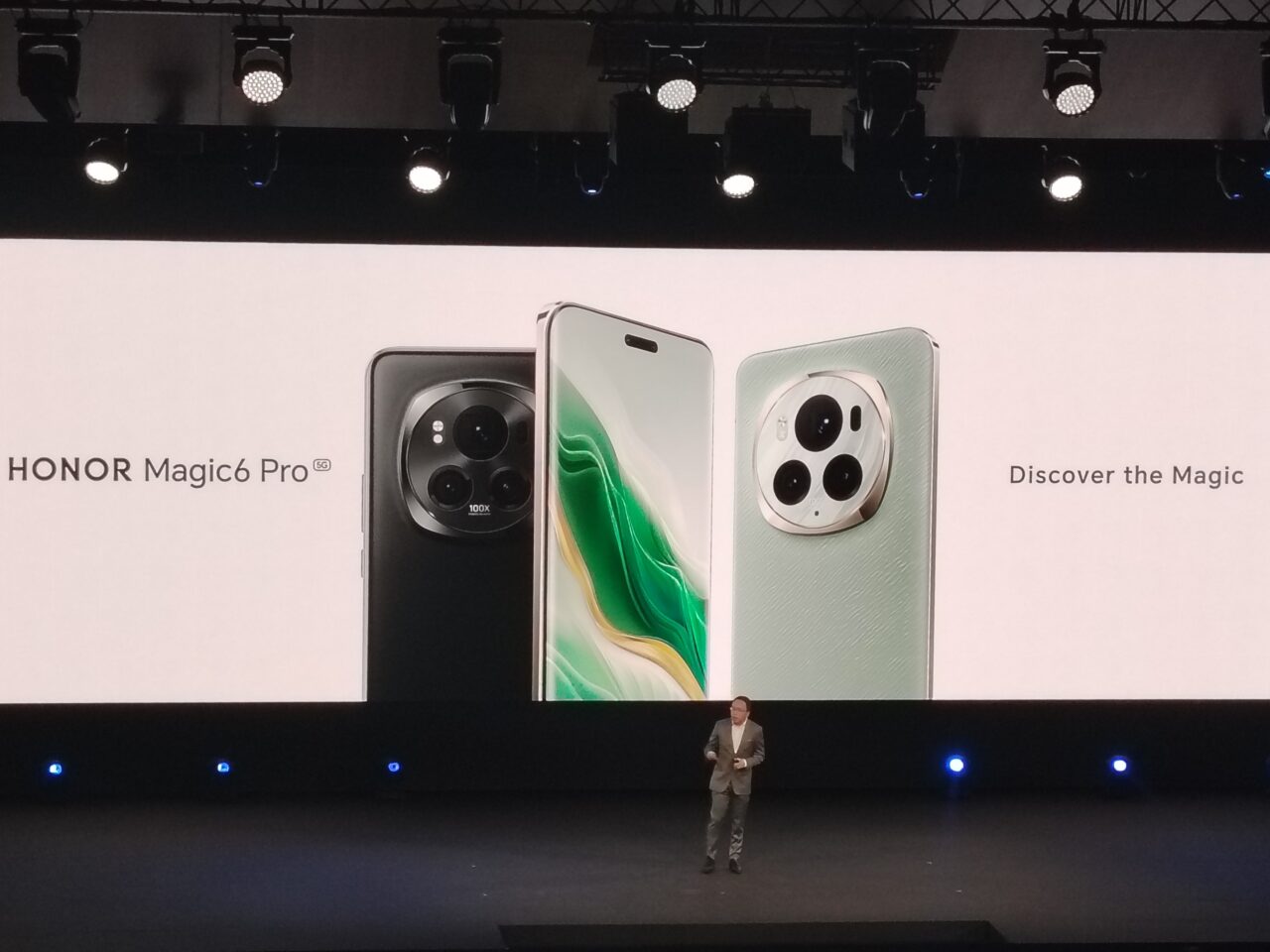 Prezentacja smartfonu HONOR Magic6 Pro na scenie z dużym ekranem wyświetlającym obrazy telefonu i hasło "Discover the Magic", w tle stojący mężczyzna w garniturze.