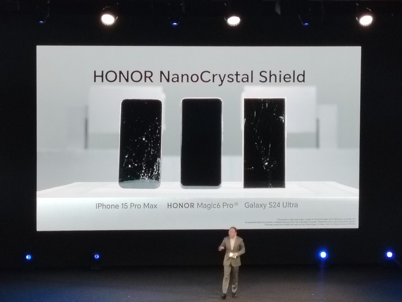 Prezentacja trzech smartfonów na scenie z uszkodzonymi ekranami, podpisane jako "iPhone 15 Pro Max", "HONOR Magic6 Pro" i "Galaxy S24 Ultra", nad nimi napis "HONOR NanoCrystal Shield", osoba stoi na scenie po prawej stronie.