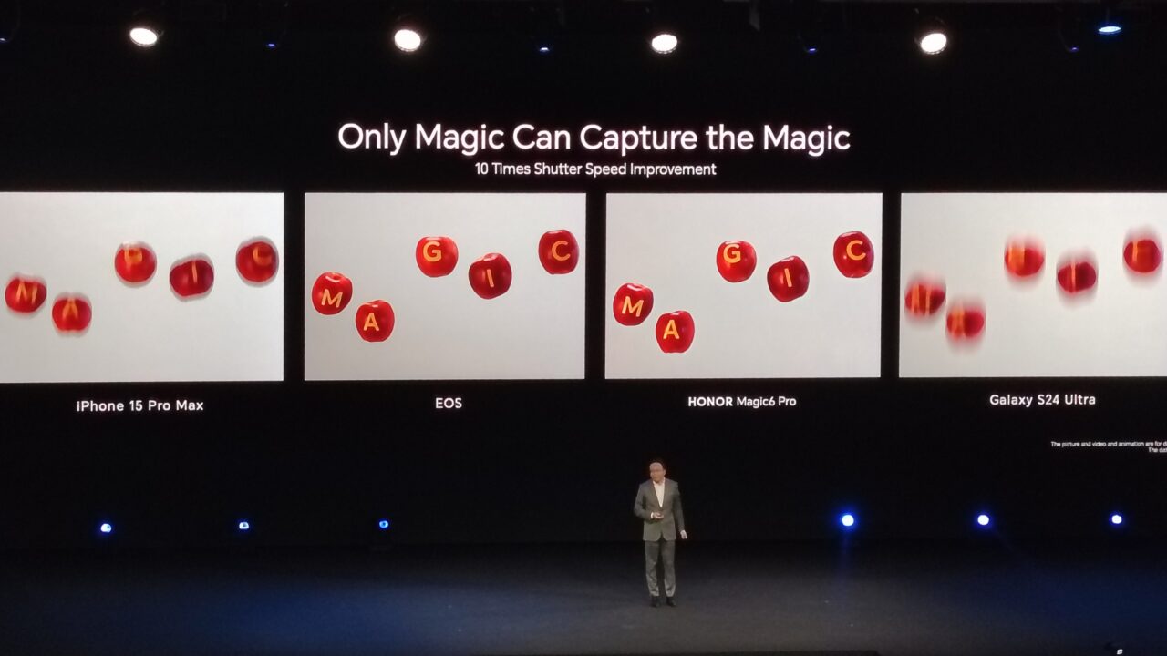 Prezentacja na scenie z mężczyzną w garniturze i czterema dużymi ekranami pokazującymi zdjęcia czerwonych jabłek z literami, które porównują jakość zdjęć wykonanych przez różne modele smartfonów: iPhone 15 Pro Max, EOS, HONOR Magic6 Pro i Galaxy S24 Ultra. Nad ekranami napis: "Only Magic Can Capture the Magic, 10 Times Shutter Speed Improvement".
