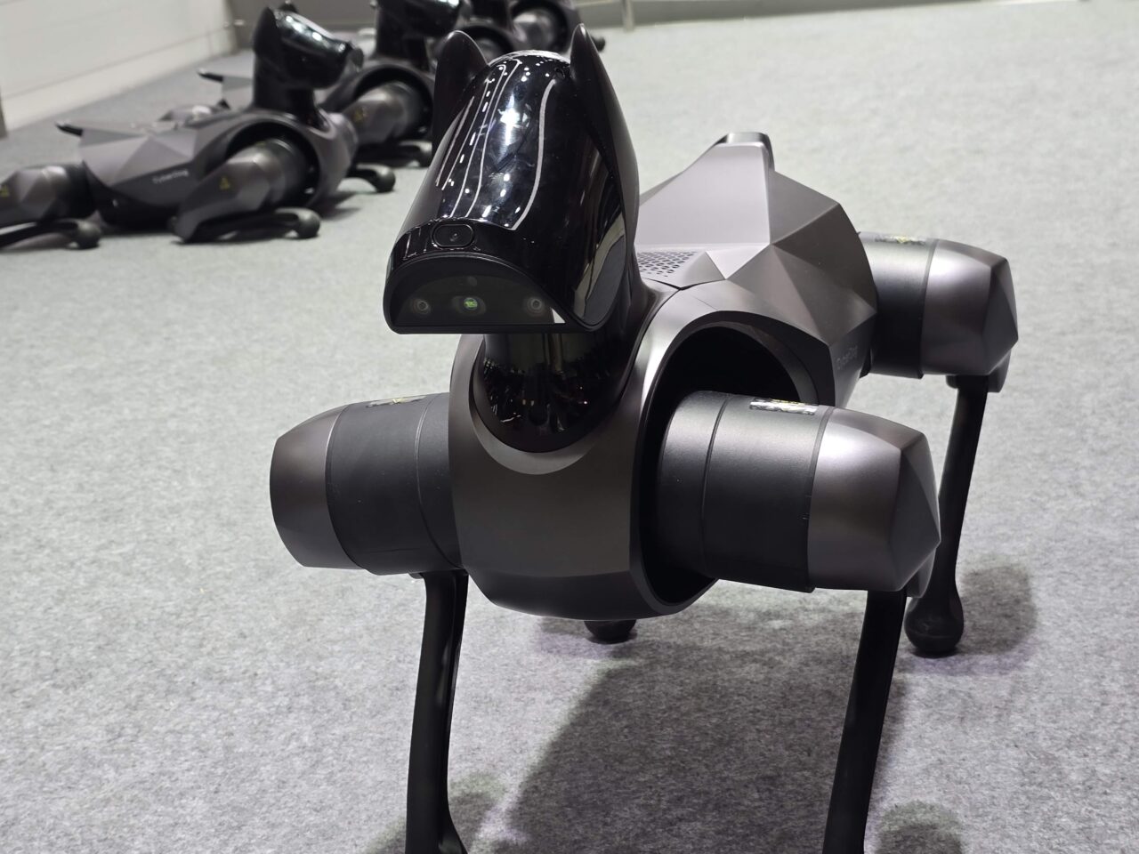 Czarno-szary robopies Xiaomi CyberDog 2 przypominający psa z "głową" wyposażoną w sensory i kamery, stojący na wystawie, z rozmazanymi robotami w tle.