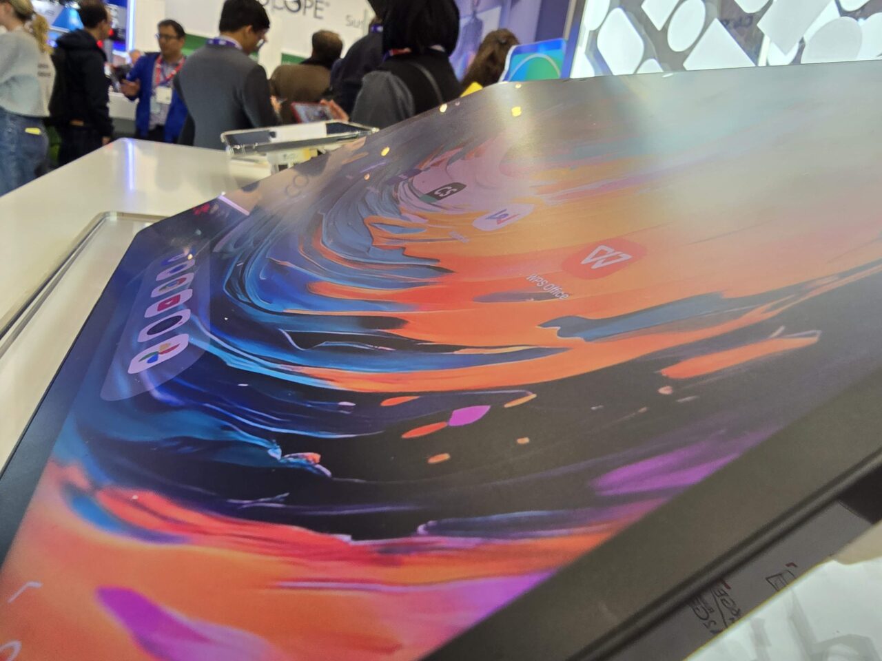 Ekrany laptopów z kolorowymi abstrakcyjnymi grafikami wystawione na targach, z tłem zamglonymi postaciami ludzi.