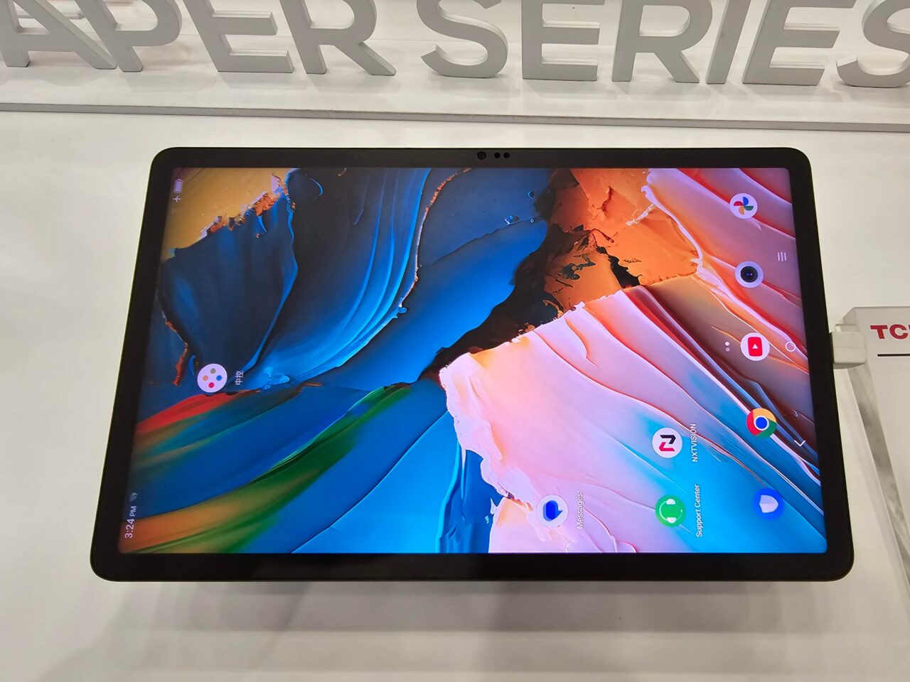 Tablet z wyświetlonym kolorowym ekranem startowym umieszczony na stojaku z napisem "PAPER SERIES" w tle.