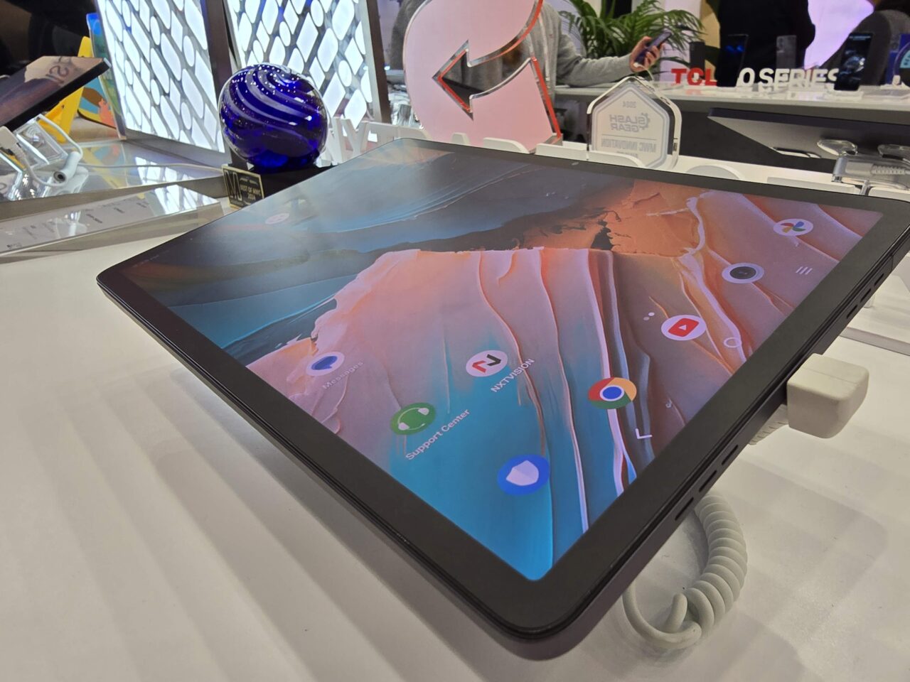 Tablet leżący na białym stole z włączonym ekranem, otoczony przez różne przedmioty, takie jak kula szklana i reklamówki.