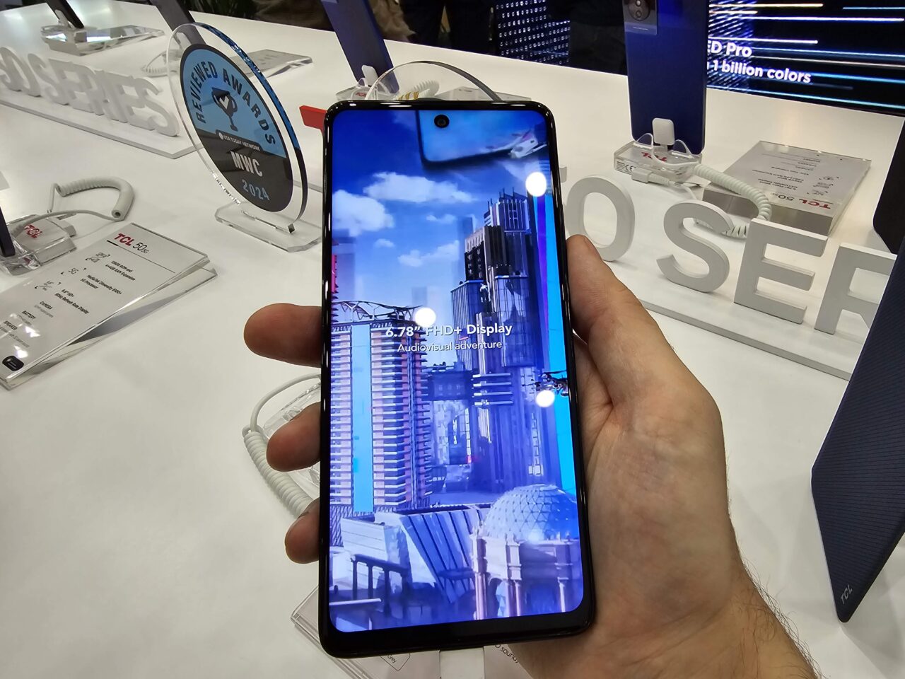 Smartfon trzymany w dłoni, wyświetlający obraz miasta na swoim ekranie, na tle wystawowego stoiska z innymi urządzeniami elektronicznymi.