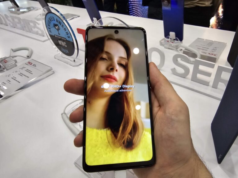 Osoba trzymająca smartphone z linii TCL 50 prezentujący ekran z aplikacją do selfi z twarzą kobiety, w tle stoisko wystawowe z nagrodami i innymi urządzeniami.