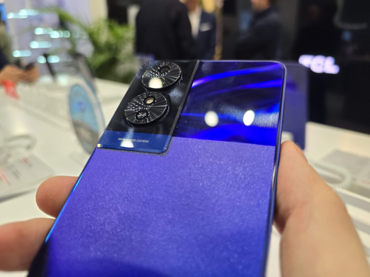 Smartfon trzymany w ręce z widocznym tylnym aparatem odblaskowym na błyszczącej niebieskiej obudowie, na nieostrych tle z elementami wystroju sklepu.