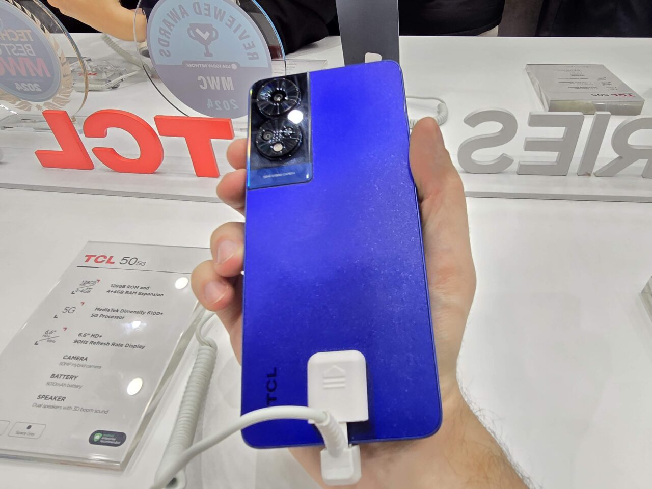 Dłoń trzyma niebieski smartfon TCL 5G z potrójnym aparatem, podłączony do bezpiecznika w sklepie.