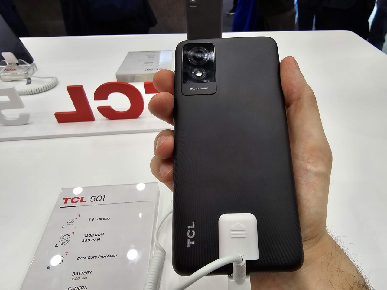 Dłoń trzymająca czarny smartfon marki TCL 501 z tyłu, z widocznym modułem aparatu i logo producenta, znajdująca się nad białą wystawą z napisem TCL i specyfikacją produktu.