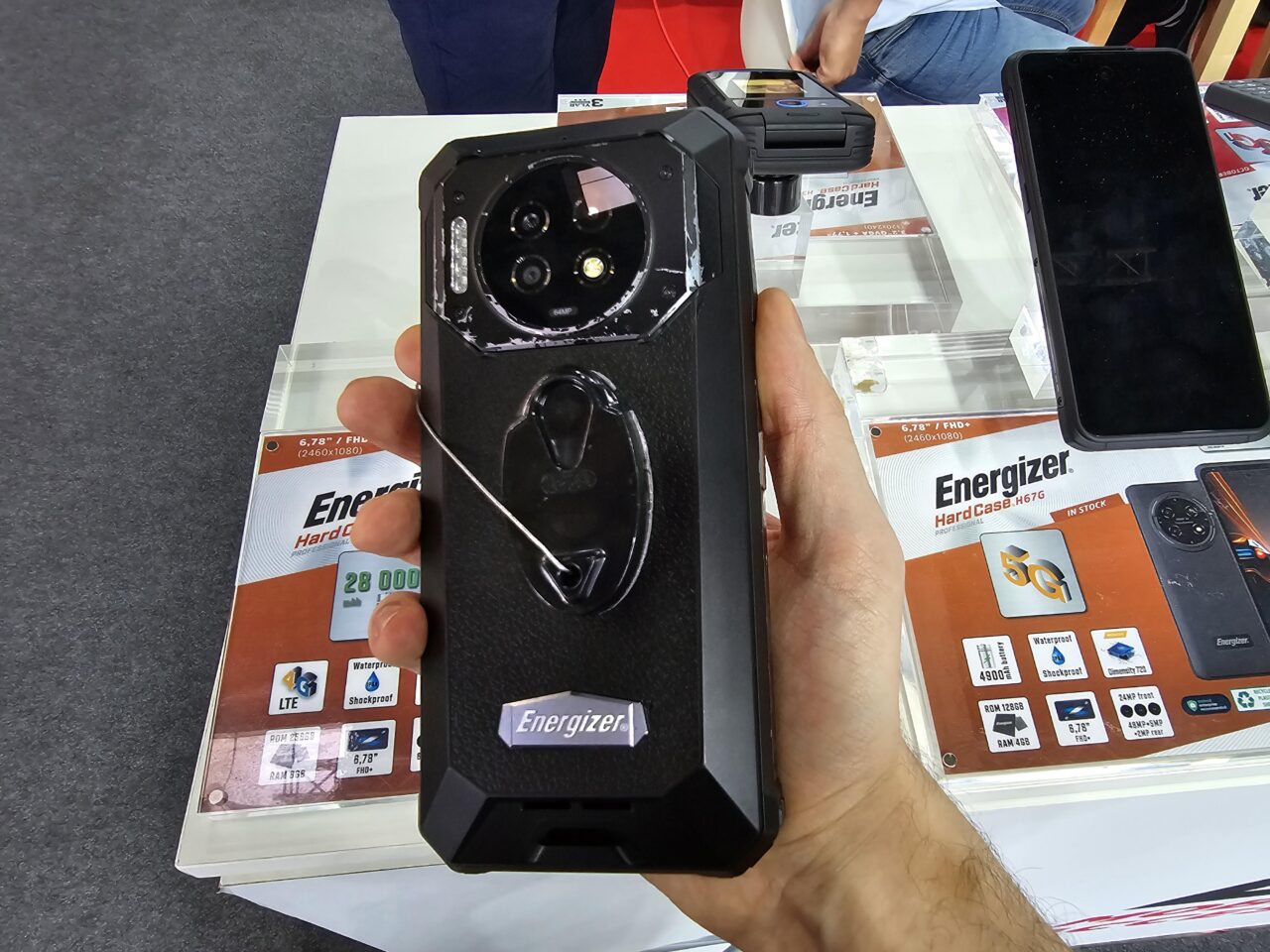 Osoba trzymająca w ręku czarny, wytrzymały smartfon Energizer Hard Case P28K z dodatkowym modułem aparatu oraz przyczepionym uchwytem na palec, na tle plakatów z informacjami technicznymi o telefonach.