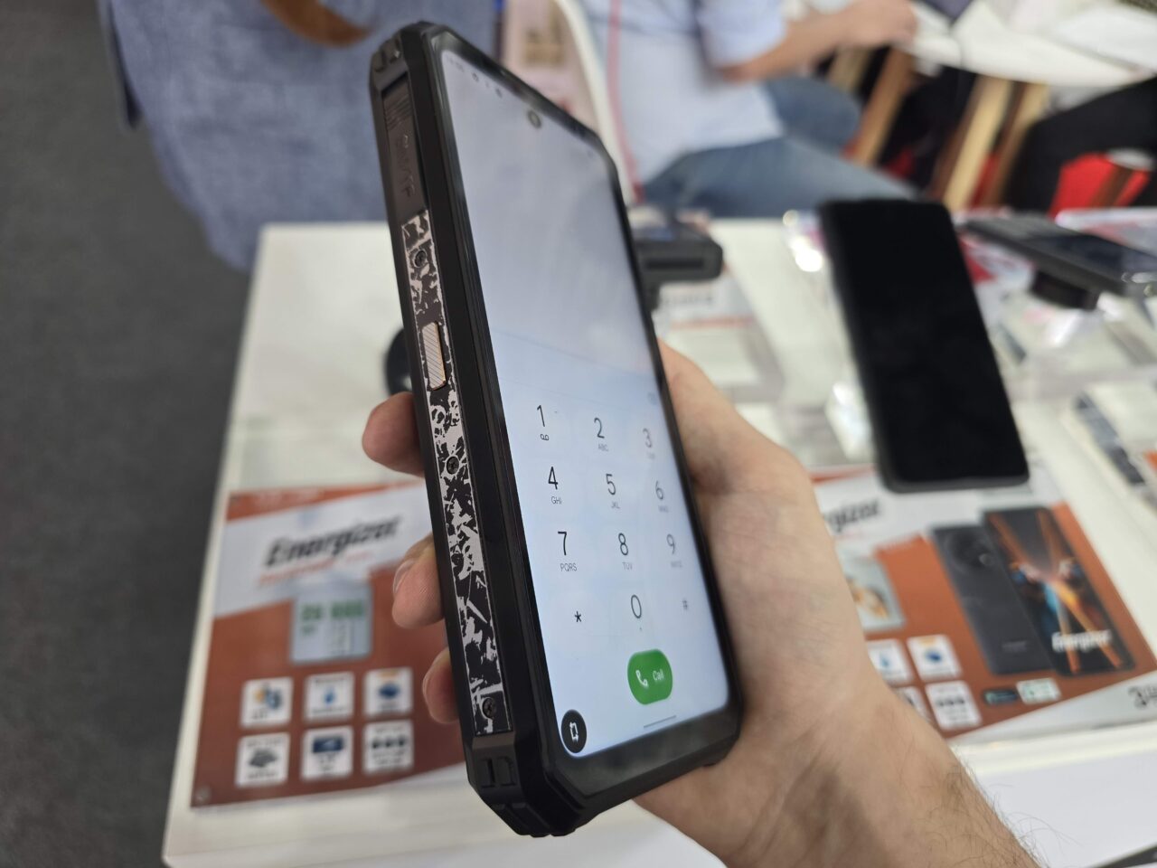 Ręka trzymająca smartfon Energizer Hard Case P28K z otwartą aplikacją do wykonywania połączeń, na ekranie widoczne są cyfry od 0 do 9, w tle rozmyty widok sklepu z elektroniką.