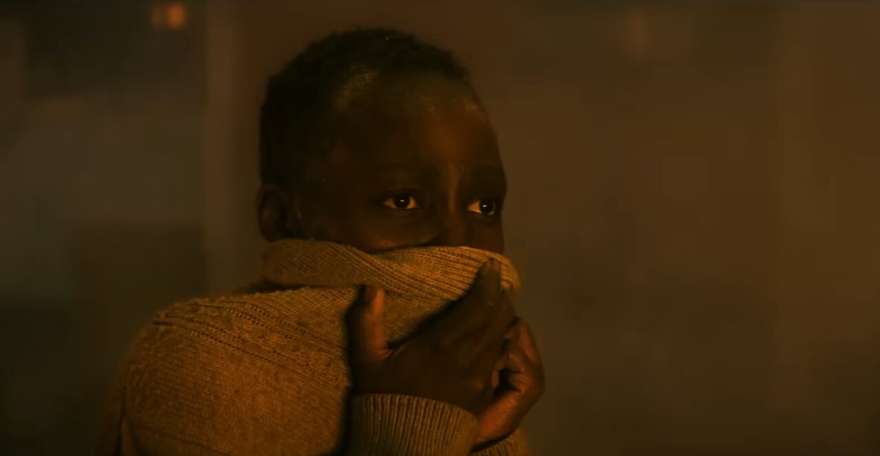Kadr z filmu Ciche Miejsce 3. Młoda osoba o ciemnej karnacji w swetrze zasłaniająca usta i nos swetrem, wydaje się wystraszona lub zaniepokojona, w pomieszczeniu z ciepłym, mrocznym oświetleniem.