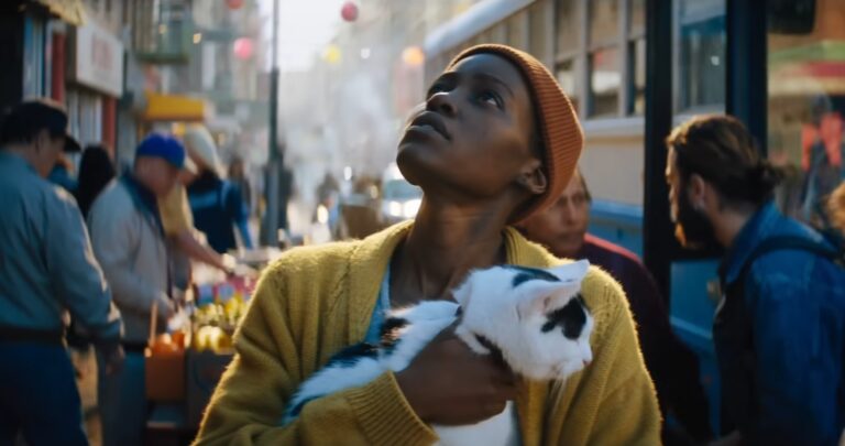 Kadr z filmu Ciche Miejsce 3. Kobieta trzymająca kota i patrząca w górę na zatłoczonej ulicy miejskiej.