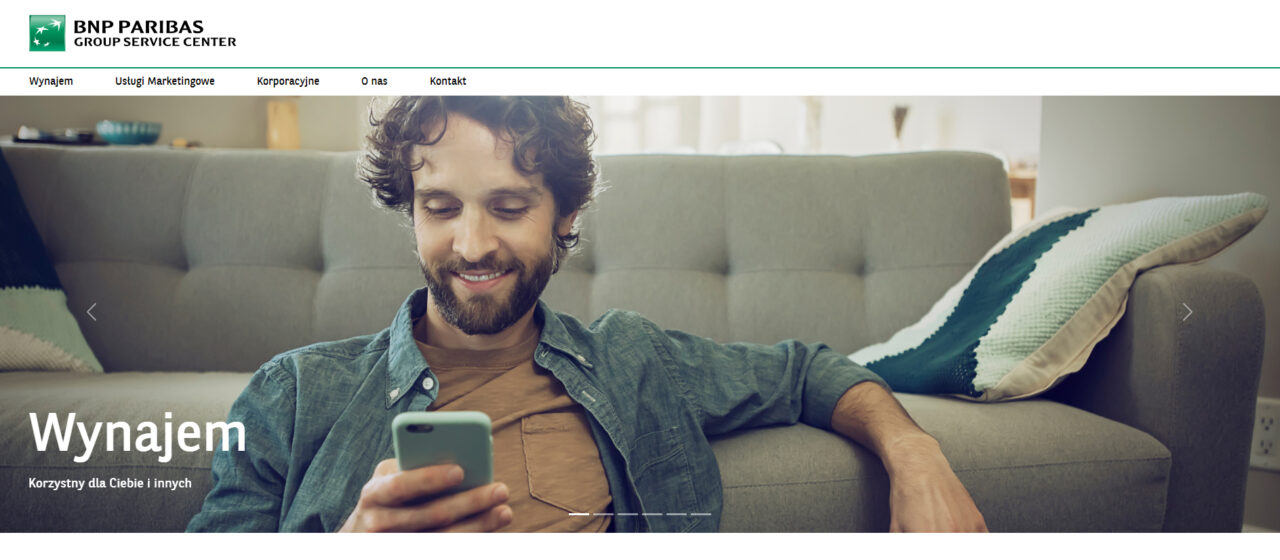 Uśmiechnięty mężczyzna siedzi na szarej sofie i patrzy na smartfon, w tle logo 'BNP PARIBAS GSC (GROUP SERVICE CENTER)', a poniżej napis 'Wynajem - Korzystny dla Ciebie i innych'.