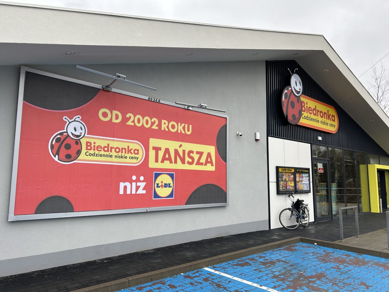 Sklep Biedronka z dużym czerwonym banerem reklamującym niskie ceny od 2002 roku w porównaniu z Lidlem, obok wejście do sklepu i zaparkowany rower.
