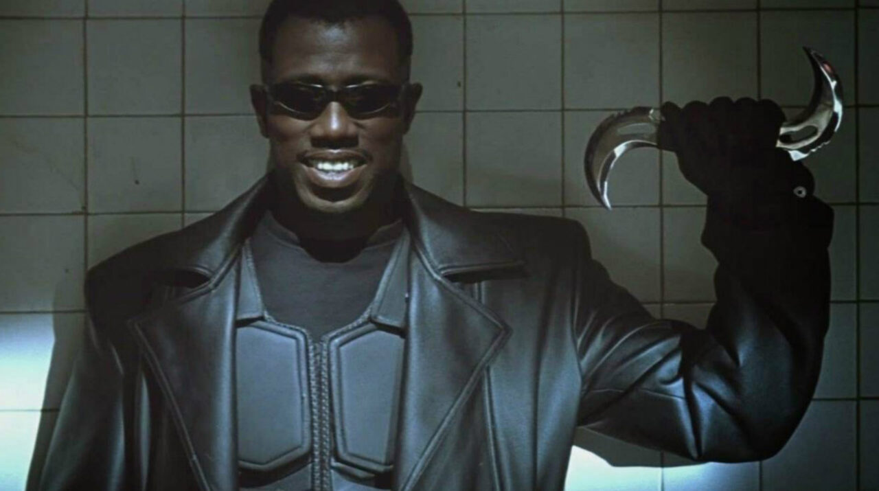 Kadr z filmu Blade. Mężczyzna w czarnej kurtce i okularach przeciwsłonecznych, śmiejący się i trzymający metalowe ostrze z haczykowatym końcem.