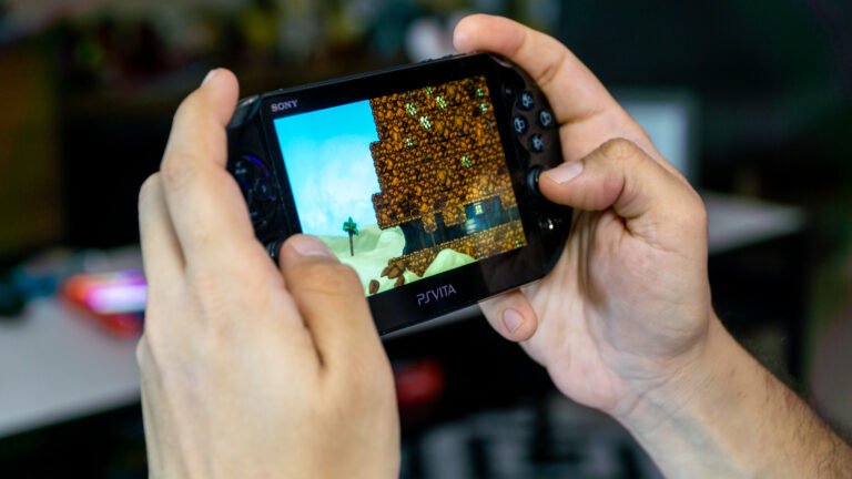 Ręce trzymające przenośną konsolę do gier Sony PlayStation Vita, na której widać grafikę z gry wideo.